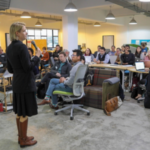 Woman speaking into a microphone to a seated audience in a modern, open-plan workspace filled with laptops and notepads.